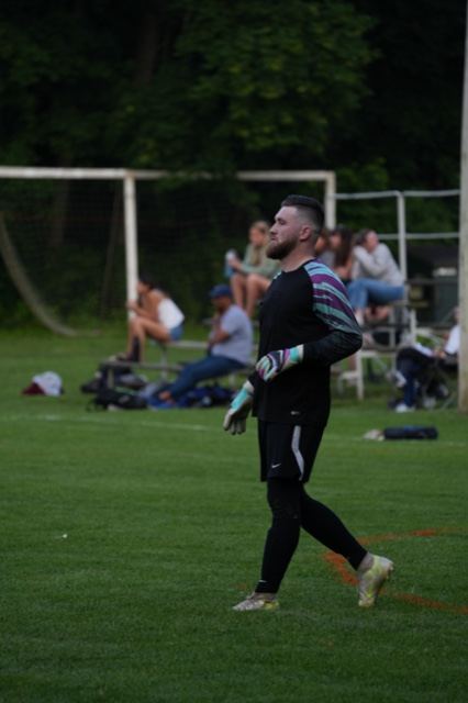 A goalkeeper walks back toward goal as spectators watch from the bleachers at Germania, June 2025