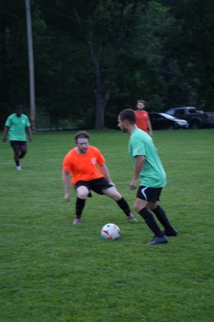 Players in orange and green battle for the ball at Germania at dusk, June 2025